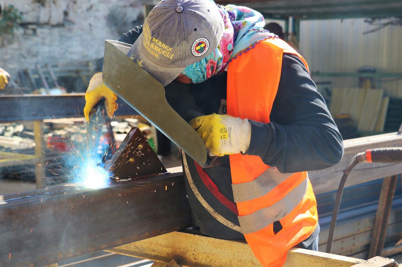 A welder wearing safety gear is welding metal beams, creating sparks outdoors.