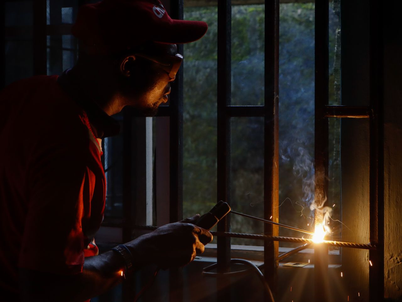 A worker in silhouette, expertly welding a metal grill with sparks flying, indoors.